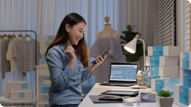 Small business owner looks at their mobile phone, sitting at a desk with product around them.