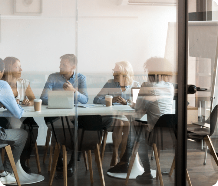 A group of people sit in a conference room behind a glass wall with the client, discussing strategy.