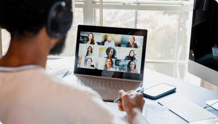 A person sits in front of a laptop with headphones on, taking notes during a virtual meeting.