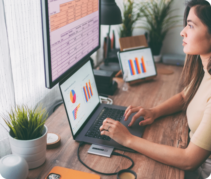 Person sits at desk with a laptop open and connected to a monitor screen showing various forms of data, charts, and graphs.