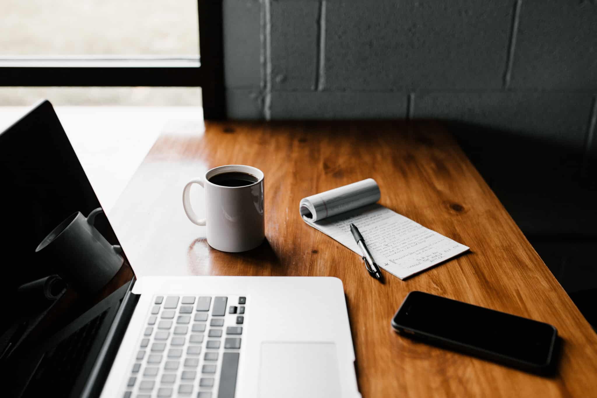 A laptop sits open on a diner table with coffee, a pen and notepad and cellphone with it.