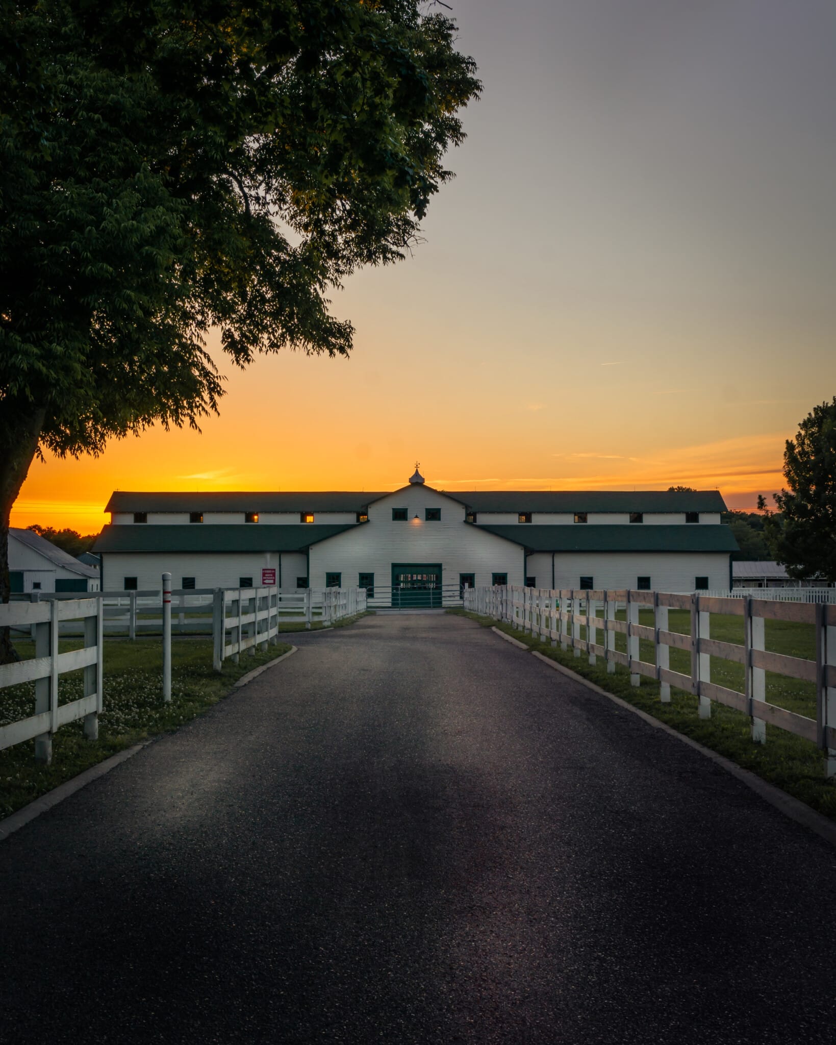White barn with white fence, sun setting in the background.