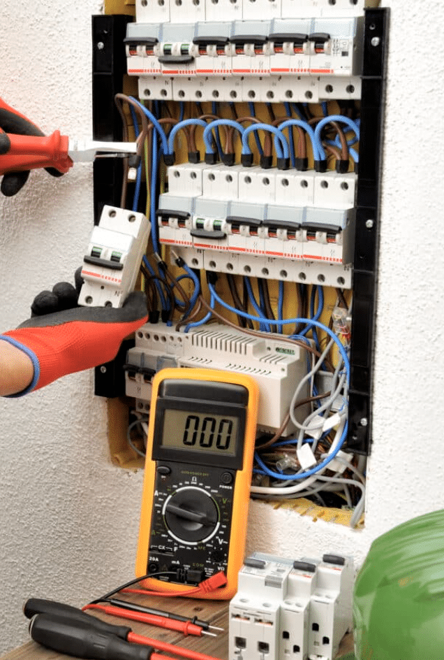 One hand holds the meter tester to wires of an electrical panel, with the meter sitting in front of the panel.