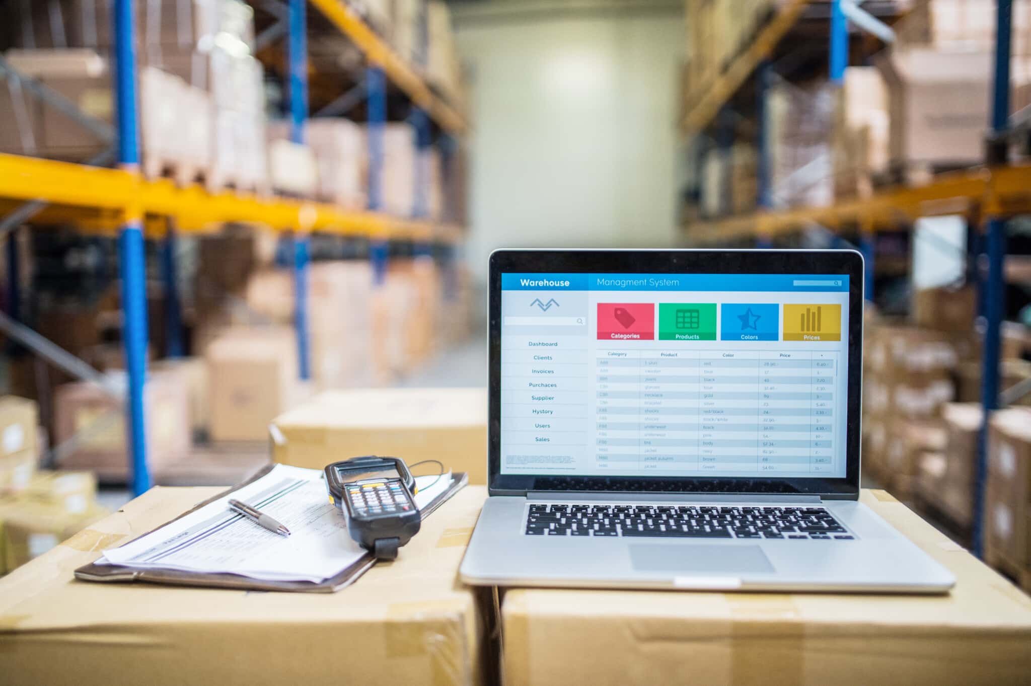 An open laptop displays warehouse inventory with a barcode scanner laying next to it and blurred shelves in the background.