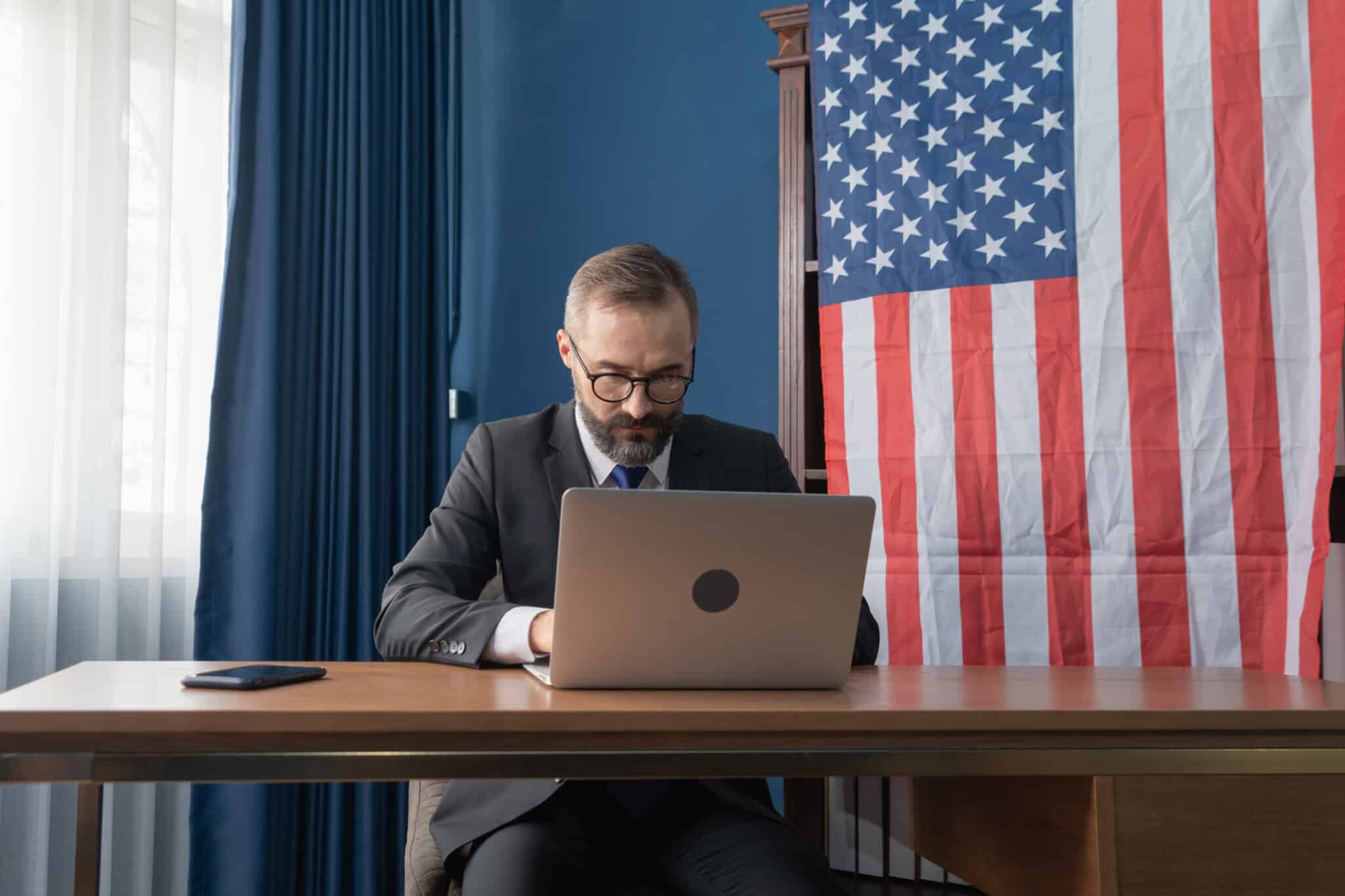 Person sits at a desk with an open laptop and United States flag behind them.