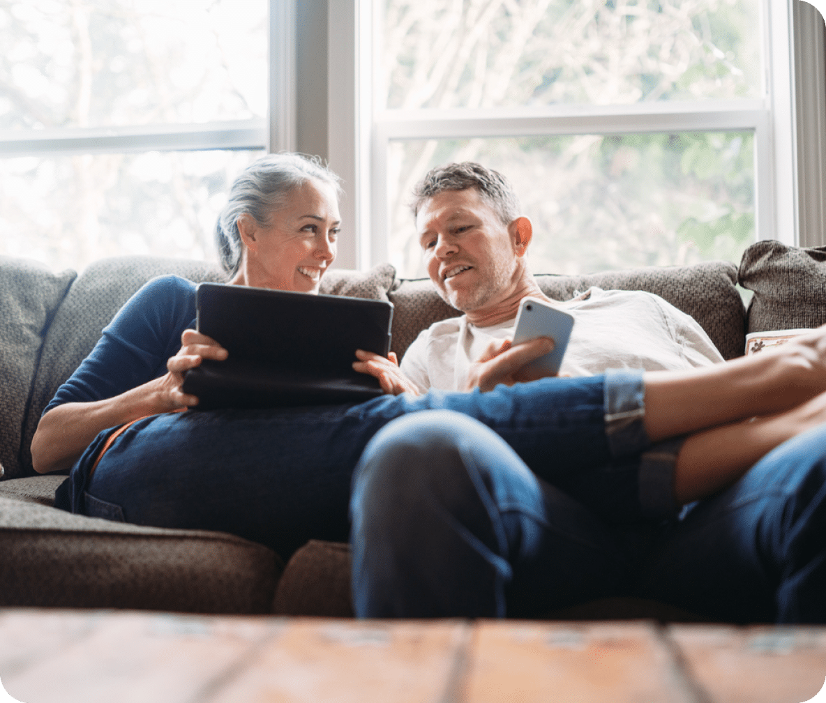 Two people sit on a couch, one holding a tablet, the other a mobile phone, smiling at each other.