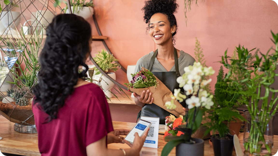 A person working at a flower shop, holds a bouquet of flowers while talking to a customer making their payment.