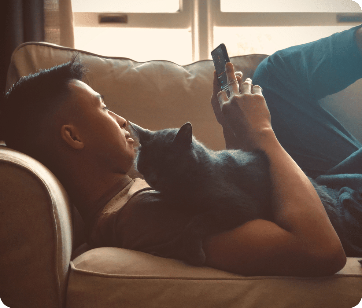 Person lays on a tan couch with a black cat on their chest as they scroll on their mobile phone.