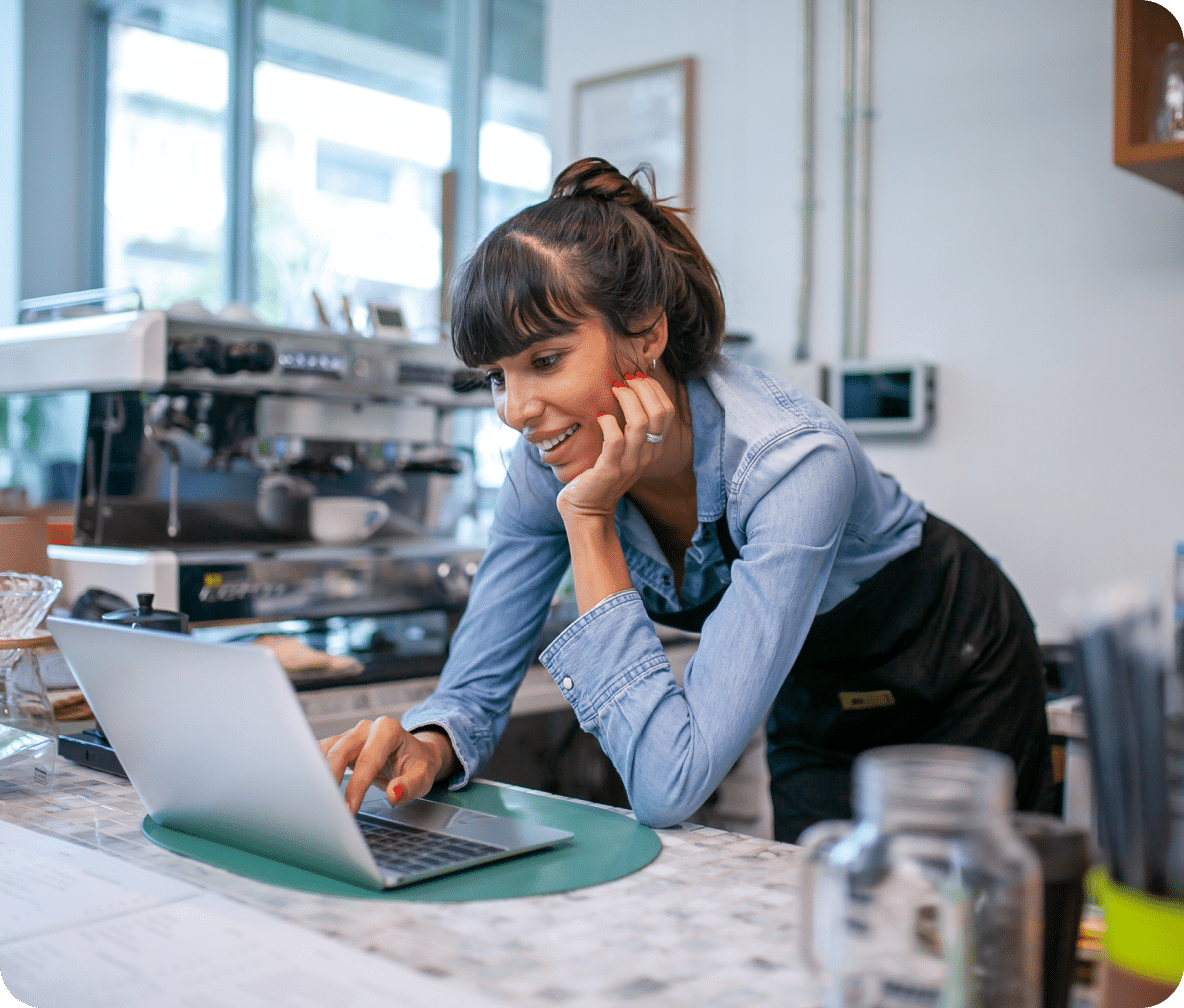 Business owner leans down on their cafe counter as they smile at their laptop.