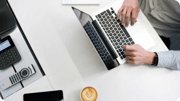 Laptop sits on a desk with a cup of coffee to the side and hands resting on the keyboard.