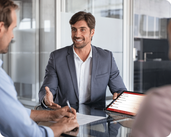 Man in a grey suit describing something with a tablet presentation in a sales business meeting while seated in a meeting room.